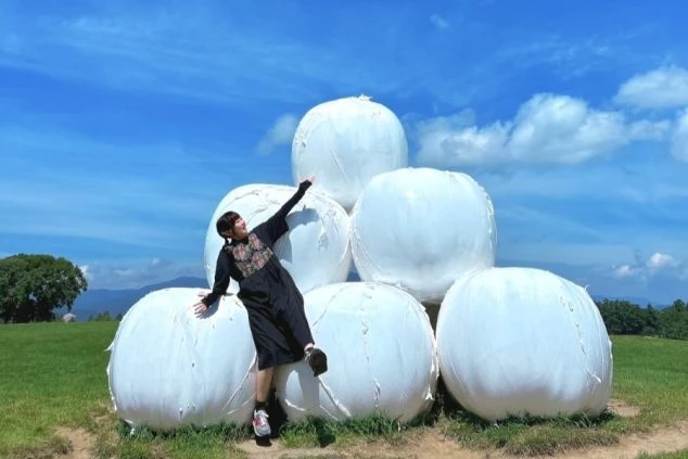 青空と緑の草原が広がる牧場で、白い丸い牧草ロールのオブジェに寄りかかってポーズをとっている若い女性ワクトリスタッフの画像。リゾートバイトの休日の過ごし方や観光を表す画像。