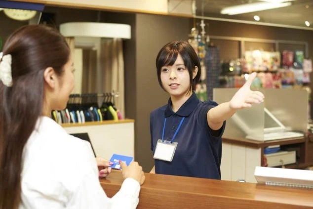 紺色のポロシャツと名札を着用した女性スタッフが、レジカウンター越しにお客様を案内している画像。売店やお土産屋でのリゾートバイトの仕事内容を表す画像。
