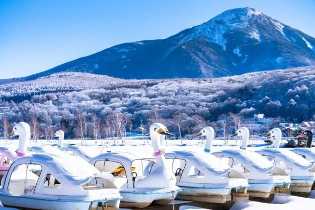 青空の下、雪化粧をした山と湖畔の風景の画像。岸辺には雪をかぶった白鳥ボートが並んでおり、冬のリゾート地の自然の風景が楽しめる。リゾートバイトの休日の過ごし方を表す画像。