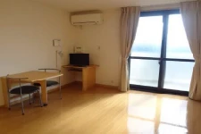 寮の居室。光沢のあるフローリングの床、大きな窓とカーテン、エアコンが設置されている。奥にはダイニングテーブルと椅子、小型のテレビ台が見える。