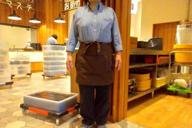 碧いシャツに茶色の腰巻気エプロン、黒スラックスにスニーカーを身につけたレストラン裏方スタッフの制服画像。