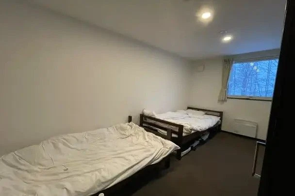 寮の居室。茶色いカーペットの部屋に、木製のベッドフレームのシングルベッドが2台置かれている。窓の外は雪景色のように白く見える。