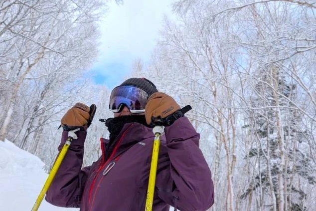 スキーウェアを着用し、ゴーグルとスキーポールを持った人物のクローズアップ画像。雪を被った木々を背景に、スキーを楽しんでいる様子が伝わる。リゾートバイトの休日の過ごし方や冬のアクティビティを表す画像。