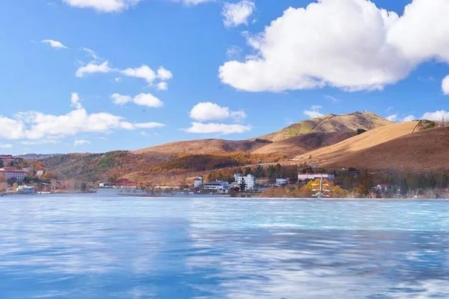青空の下、湖畔にリゾートホテルが建ち並び、紅葉に色づいた山々が広がる雄大な風景の画像。リゾートバイトの休日の過ごし方や観光を表す画像。