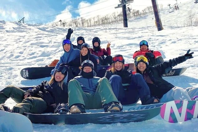 スキー場の雪山で、スノーボードを持った若い男女の大人数グループが笑顔でカメラにポーズをとっている画像。冬のアクティビティを楽しむリゾートバイト仲間の楽しそうな雰囲気が伝わるワクトリオリジナル画像。