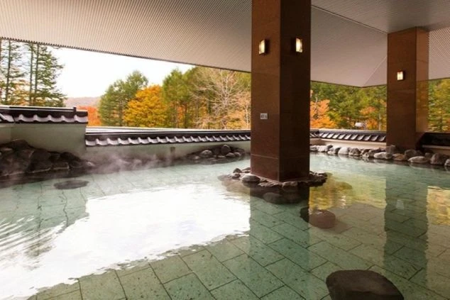 紅葉に色づいた木々が見える露天風呂の画像。湯気が立ち上る広い浴槽は石で囲まれ、和風の屋根が付いている。リゾートバイトの寮やホテルの福利厚生として利用できる、温泉の画像。