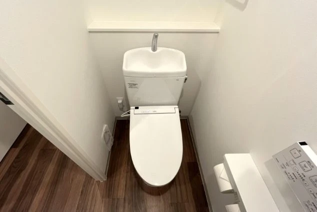 手洗いボウル付きの洋式トイレ。温水洗浄便座が完備され、背後の壁にはちょっとした小物が置ける棚がある。床はダークブラウンの木目調で落ち着いた雰囲気。