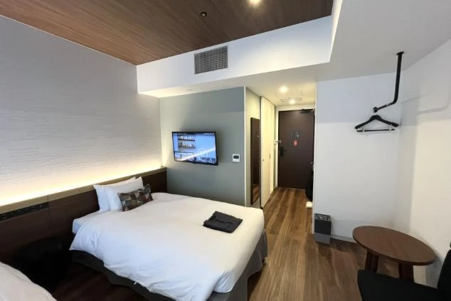 木目調の床とグレーの壁がおしゃれな洋室。大きなダブルベッドが中央にあり、壁掛けテレビ、小さな丸テーブル、オープンタイプのハンガーラックが備わったモダンな個室。