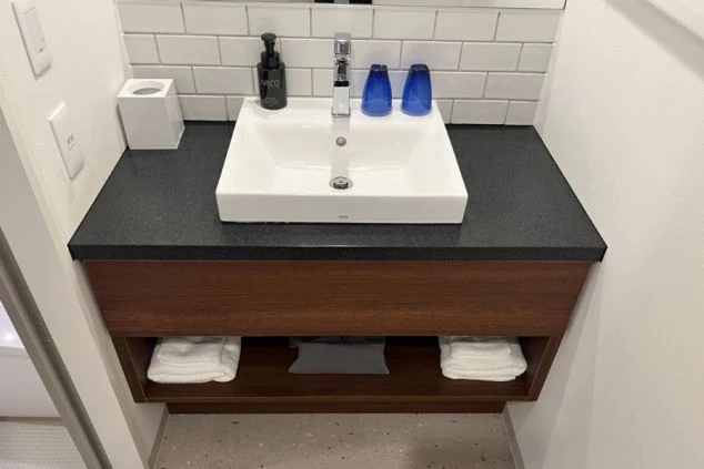 黒い天板に白いスクエア型の洗面ボウルが映える洗面台。下部の棚には予備のタオルが綺麗に畳んで置かれ、壁は白いタイル調のデザインで清潔感がある。