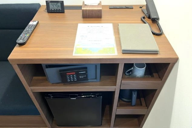 機能的な収納デスク。天板には時計や電話があり、棚には小型の金庫、電気ケトル、マグカップ、さらに下段には小型冷蔵庫がコンパクトに収納されている。