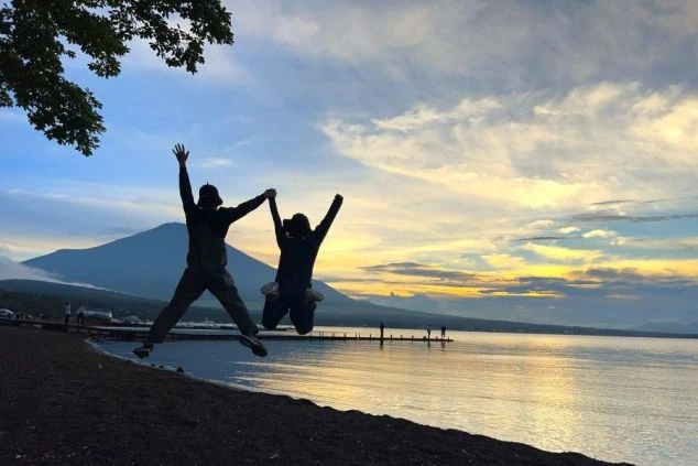夕焼けの湖畔で、手を取り合ってジャンプするリゾートバイトの仲間のシルエット写真。仕事の合間や休日に、こんな絶景の中で非日常的な体験を楽しめる魅力を伝えます。