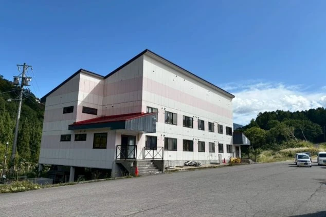 山間に建つリゾートバイト寮の外観。ピンクと白のツートンカラーの建物で、スキー場や温泉地に近く、住み込みで働くための寮。