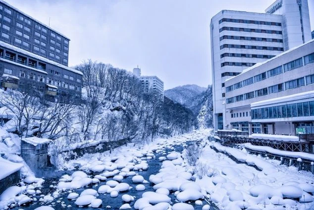 雪景色に覆われた温泉街の風景の画像。雪が積もった川の両岸にホテルが立ち並び、冬のリゾート地の景色が広がる。リゾートバイトの休日の過ごし方や観光を表す画像。