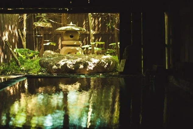 日差しが差し込む露天風呂のような温泉の画像。石灯籠や岩、緑の植物が配置され、落ち着いた雰囲気が漂う。リゾートバイトの寮やホテルの福利厚生として利用できる、温泉の画像。