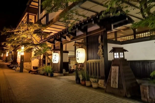 夜の温泉旅館のエントランスの画像。木造の建物が提灯と木々のライトアップで温かく照らされ、風情ある街並みが写っている。リゾートバイトの仕事場となる旅館の外観の画像。