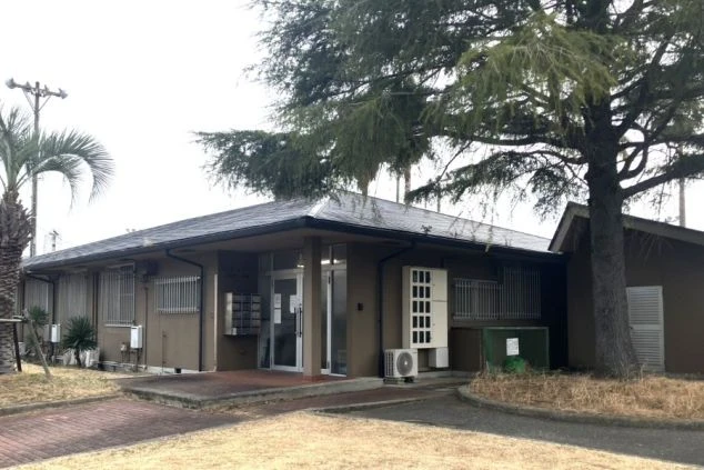 豊かな緑に囲まれたスタッフ寮の外観画像。落ち着いたブラウンカラーの平屋建築で、集合ポストや玄関が整えられた安心の住み込み拠点。