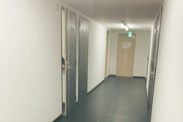 リゾートバイトの寮の廊下の画像。白い壁とドアが続き、非常口のサインが見える。共同生活の場としての寮の内部を示す。