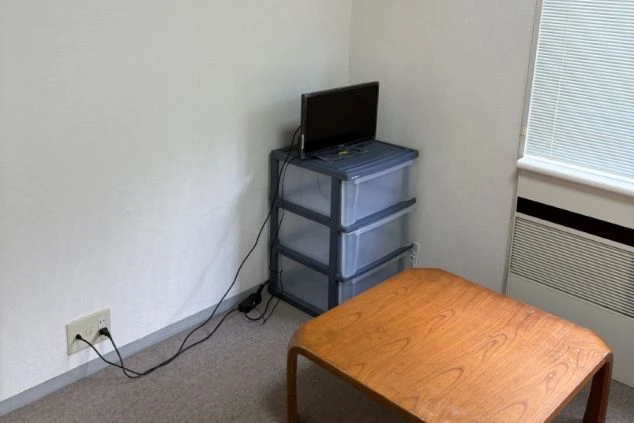 小さなテレビと3段のプラスチック収納ケースが置かれた寮の室内の画像。