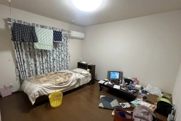 寮の居室。フローリングの床にシングルベッドとローテーブル、ブラウン管テレビが置かれ、窓際には洗濯物が干されたカーテンとエアコンがある。