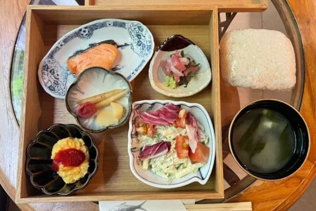  焼き鮭、刺身、サラダ、卵料理、漬物、ご飯、味噌汁が木製の箱にきれいに盛り付けられた和食の食事。