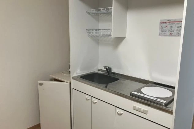 寮のキッチン。シンク、コンロ、冷蔵庫、吊り戸棚、網棚が映る。