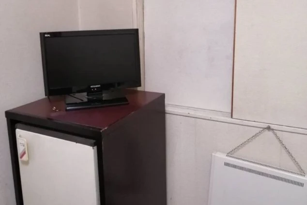 寮の個室。テレビと冷蔵庫がセットの収納棚、壁に暖房器具が映る。