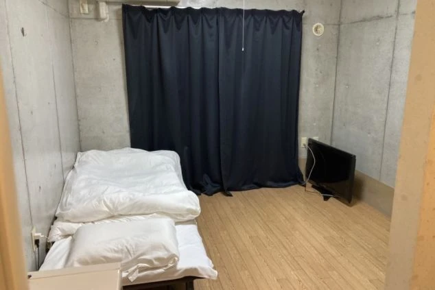 寮の居室。コンクリート打ちっぱなし風の壁と木目調の床。窓際には黒い遮光カーテンがかけられ、ベッドと小型テレビが置かれている。