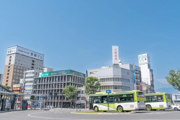 山梨県甲府駅前の街並みの画像。青空の下、ビルが立ち並び、緑色のバスが停車している。リゾートバイトの仕事場や周辺環境を表す都会の風景の画像。