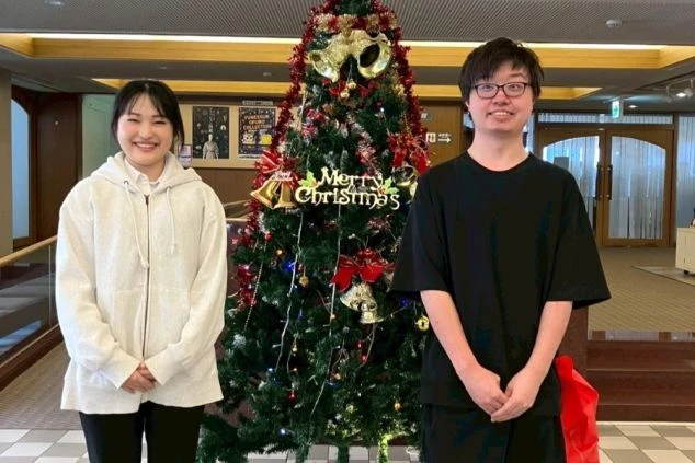 クリスマスツリーを挟んで並ぶ、白いパーカーの女性と黒いTシャツの男性。ホテルのロビーのような場所で笑顔を見せている。