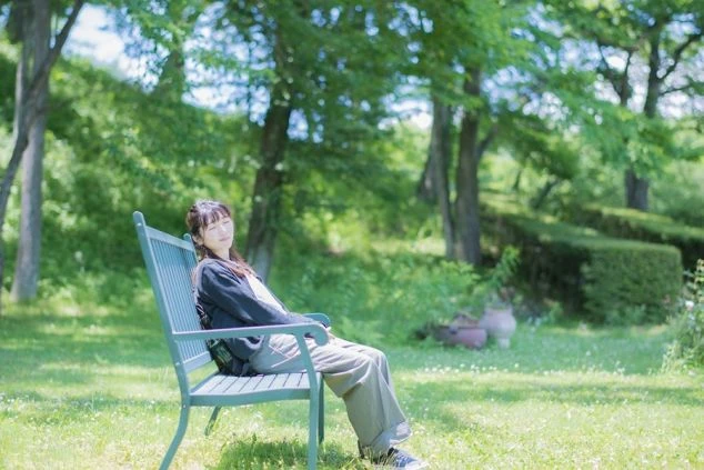 緑の木々と芝生が広がる公園のベンチで、若い女性が目を閉じて座りリラックスしている画像。穏やかな日差しが差し込む自然の中で、リゾートバイトの休日を過ごしている様子が伝わる画像。