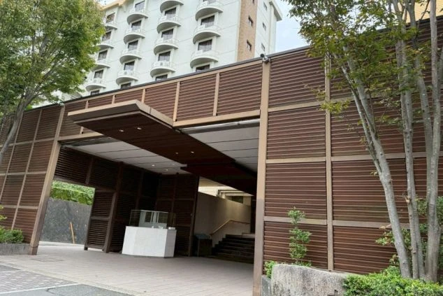 木目調のルーバーで覆われたモダンな車寄せの先に、屋内へ続く階段が見える。奥には客室のバルコニーが連なる高層棟の一部が写っている。