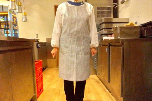 白いスモックに黒いパンツ、その上におあと白の細かいストライプ柄のエプロンを着用した調理補助スタッフの制服画像。