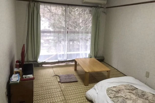 寮の個室。畳の床に布団、ローテーブル、テレビ、エアコン、窓にカーテンが映る。