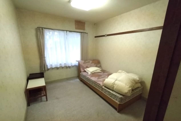 寮の個室。ベッド、サイドテーブル、窓にカーテン、壁にハンガーポールが映る。