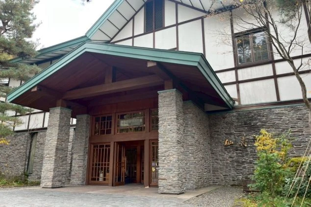 大きな石積みの柱と壁が特徴的で、木目調の玄関扉と、緑色の屋根の庇がある。上部は白壁に木の梁がデザインされた洋風の構造になっている。