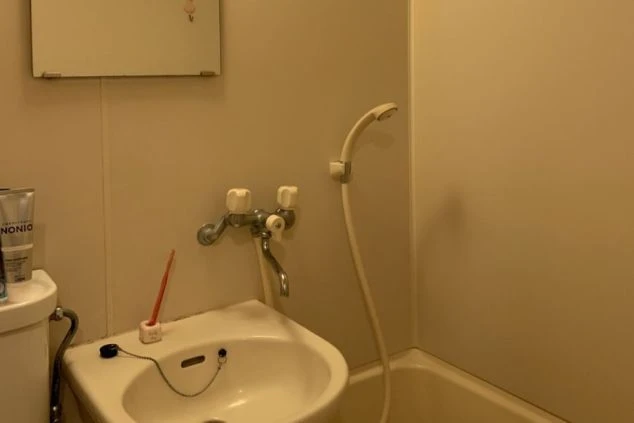 バスルームの内部を写した画像。薄いベージュ色の壁があり、左下には白い便座が見えます。壁にはトイレットペーパーホルダーらしきものが設置されており、全体的に清潔で整頓された印象です。