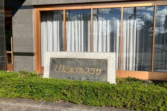 石の看板（サインボード）。手前には刈り込まれた植え込みがあり、奥には窓ガラスと白いカーテンのある建物のエントランスが見える。