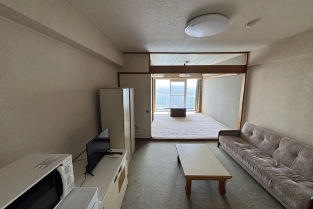 寮の共有リビングまたは居室。カーペット敷きの部屋にソファとローテーブルがあり、奥の部屋とは襖のような間仕切りで区切られている。テレビと電子レンジ、窓の外に海が見える。
