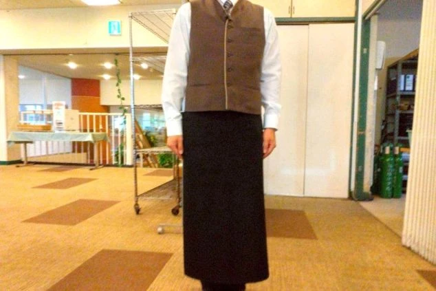 白い長袖シャツに茶色のベスト、黒いロングエプロンを着用した男性スタッフの全身画像。リゾートバイトの仕事内容を表す、高原ホテルのマルチ業務スタッフの制服の画像。