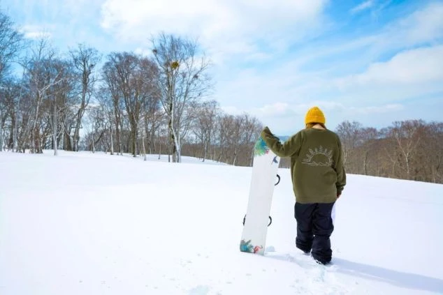 雪原をスノーボードを脇に抱えて歩く女性ワクトリスタッフの後ろ姿の画像。青空と木々の雪景色が広がり、冬のアクティビティを楽しむリゾートバイトの休日を表す画像。