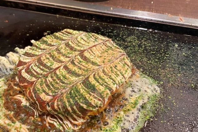 鉄板の上で焼かれている、熱々のお好み焼き。マヨネーズとソース、青のりがかけられた、食欲をそそる大阪グルメの代表格です。