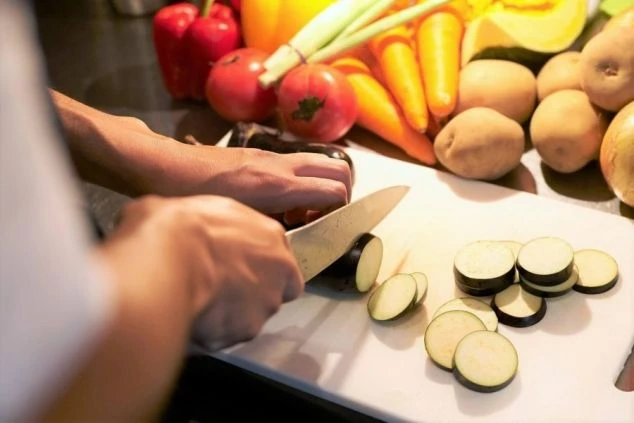 まな板の上でナスをスライスする手元のクローズアップと、周りに置かれた色鮮やかな野菜。調理補助の仕事内容をイメージさせる画像。