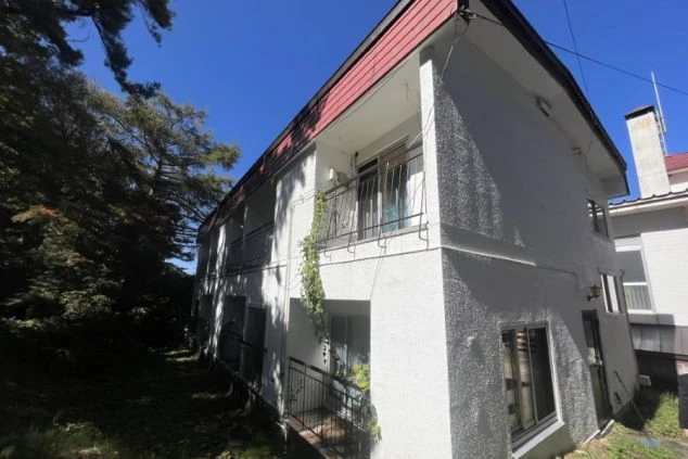 快晴の青空の下、白い壁と赤い屋根が特徴的な2階建てのリゾートバイト寮の外観の画像。バルコニーには黒い手すりがあり、蔦が絡まっている。リゾートバイトの寮の外観の画像。