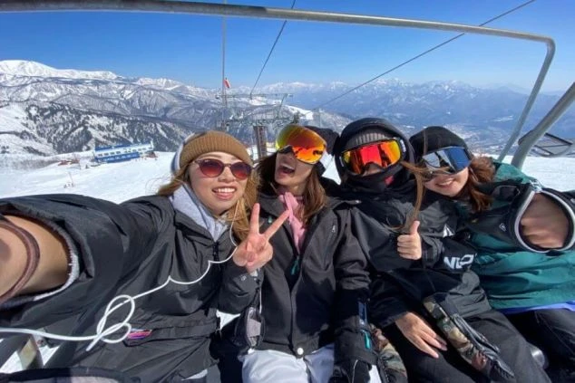 スキー場のリフトの上で、仲の良い女性4人組が楽しそうに記念撮影（セルフィー）。後ろには、雪化粧をした雄大な山々の絶景が広がっています。