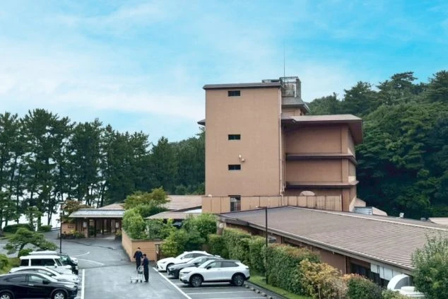 緑に囲まれた和風の建物。茶色い壁が特徴的で、手前には駐車場と入口が見えます。