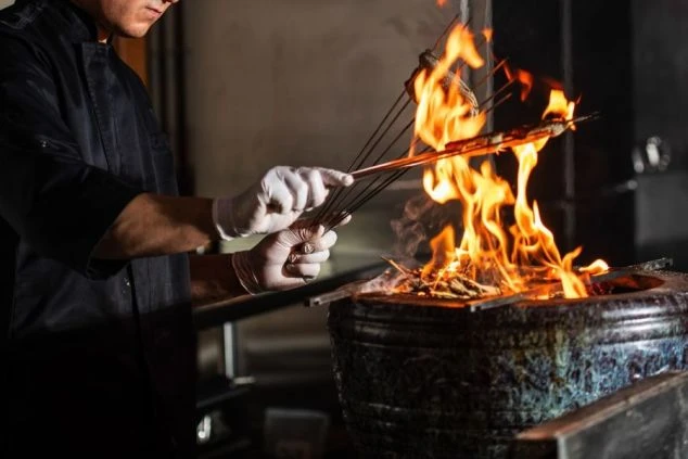 旅館やホテルの厨房。調理スタッフが炎で魚を焼いている様子。調理補助・キッチンスタッフの仕事風景。