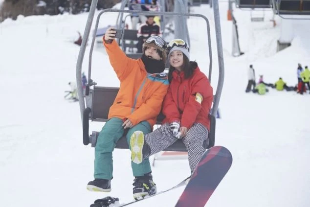 スキー場のリフトに乗って自撮りを楽しむ若い男女のスノーボーダー。リゾートバイトのリフト係として働きながら、雪山を満喫。