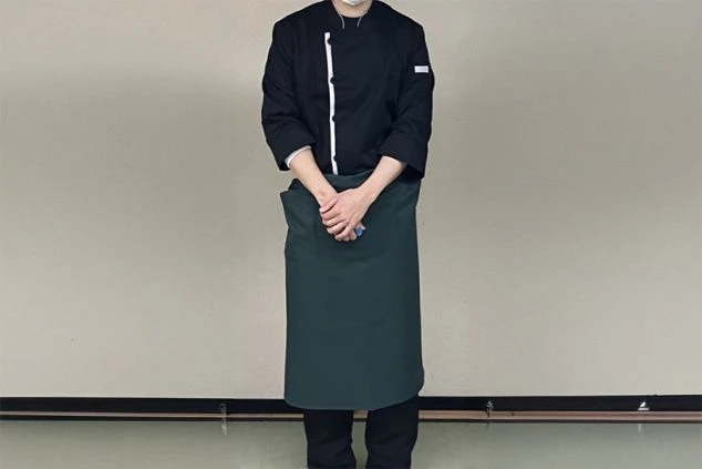 黒いコックコートと濃い緑色のロングエプロンを着用した男性が、正面を向いて立っている様子。