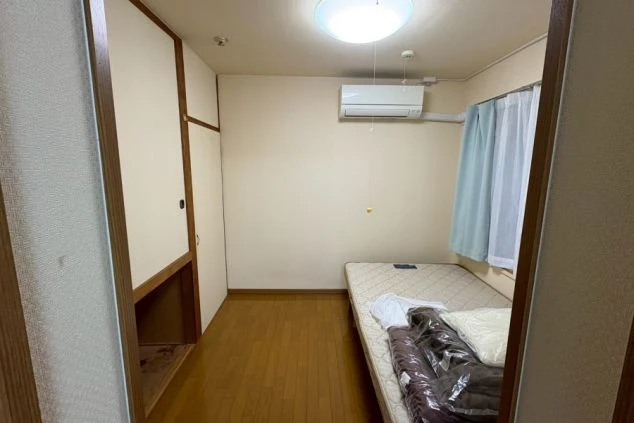 寮の個室。木目調の床にマットレスが敷かれ、奥には押入れ(またはクローゼット)とエアコン、窓が見える。