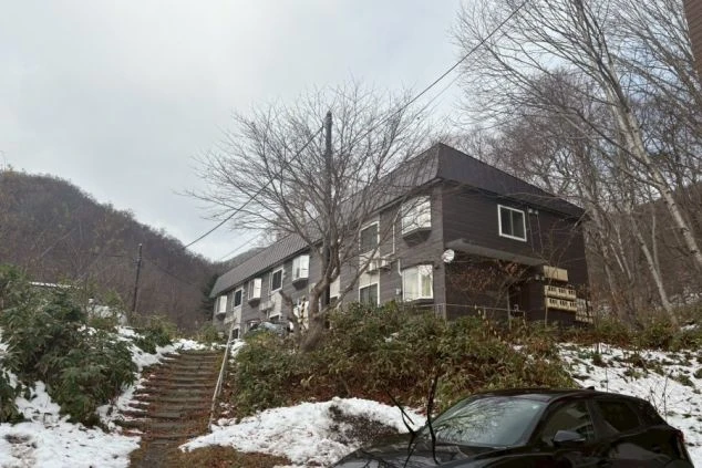 寮の外観。雪が積もった山中に建つ、濃い茶色の2階建ての建物。敷地内には階段があり、周囲は木々に囲まれている。